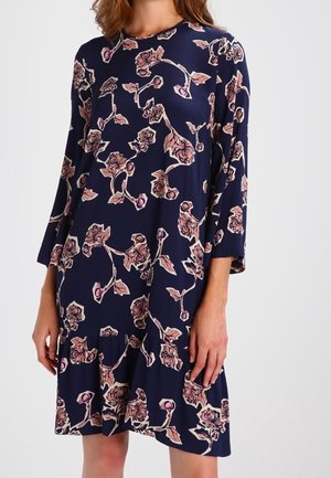 Laivastonsininen polvipituinen mekko, jossa on vaaleanpunainen kukkakuvio, pitkät hihat ja löysä röyhelöinen helma.