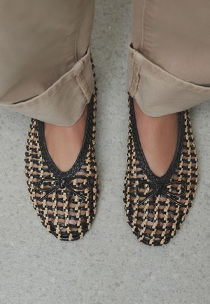 Pieds portant des ballerines tissées noires et couleur naturelle avec petits nœuds, associés à un pantalon beige sur un sol gris.