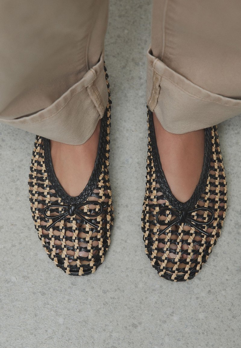 Pieds portant des ballerines tissées noires et couleur naturelle avec petits nœuds, associés à un pantalon beige sur un sol gris.