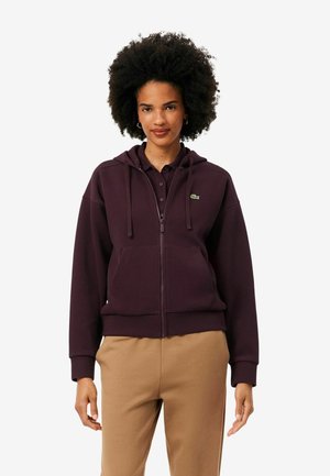 Femme aux cheveux bouclés portant un sweat à capuche violet foncé zippé et un pantalon beige, debout devant un fond uni.