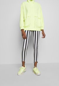 Persona vistiendo una chaqueta amarilla neón, pantalones de rayas verticales en blanco y negro, y zapatillas amarillas neón a juego, de pie frente a un fondo liso.