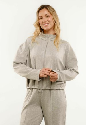 Donna sorridente con capelli biondi ondulati che indossa una giacca grigio chiaro con cerniera e pantaloni abbinati, in piedi davanti a uno sfondo bianco uniforme.