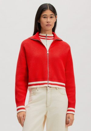 Jonge vrouw die een rode gebreide trui met rits draagt, met witte strepen aan de manchetten en zoom, gecombineerd met lichtbeige broek met hoge taille.