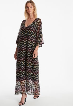 Robe longue et transparente avec un motif en chevrons en noir, rose, or et crème. Présente un décolleté en V profond et des manches larges.