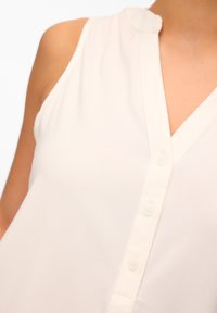 Blusa senza maniche beige chiaro con scollo a V, in morbido tessuto, chiusura con bottoni e dettaglio del colletto rotondo. Texture liscia con un design semplice.