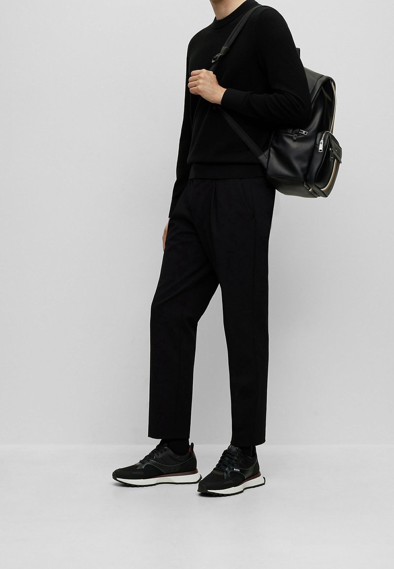 Schwarzer Pullover kombiniert mit schwarzen, plissierten Hosen und dunklen Sneakers. Ein schwarzer Rucksack mit einem Reißverschluss wird über eine Schulter getragen.