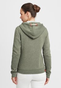 Green sweatshirt with a hood, featuring a textured fabric, drawstring detail with leather accents, and ribbed cuffs. White pants visible.