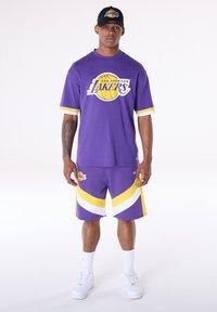 Lila Los Angeles Lakers mez fehér és sárga díszítéssel, hozzáillő rövidnadrággal, amely fehér csíkokat és logót tartalmaz. Fehér tornacipő és sapka.