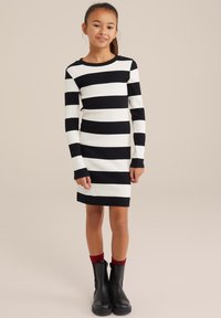 Schwarz-weiß gestreiftes Langarmkleid mit gerippter Textur, das einen runden Ausschnitt und eine taillierte Silhouette aufweist. Schwarze Knöchelstiefel mit roten Socken.