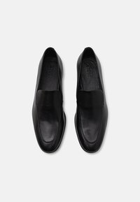 Black leather slip-on shoes with a smooth texture, elongated shape, and minimal detailing. Interior features soft lining with logo.