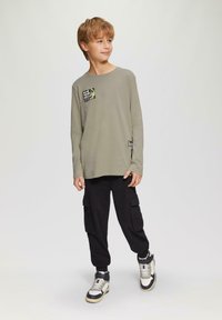 Olivenfarbendes Langarm-T-Shirt mit einer grafischen Tasche, kombiniert mit schwarzen Cargo-Hosen und hochhackigen Sneakern mit einem schwarz-weißen Design.