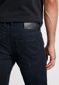 Tmavě modré džínové kalhoty s přiléhavým střihem, vybavené koženou štítkem Diesel na pásku a dvěma zadními kapsami s jemnými detaily prošití.