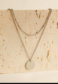 Collana a strati in argento con due catene e un ciondolo tondo e liscio. Lo sfondo è costituito da una pietra chiara e texture.