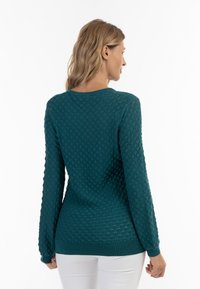 Maglione teal con un motivo a diamante testurizzato, maniche lunghe e orlo a coste. Indossato con pantaloni bianchi, che mettono in evidenza una silhouette aderente.