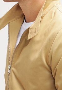 Lys beige bomberjakke med lynlås foran, spidse kraver og glat tekstur. Accentueret med metallisk lynlåss hardware.