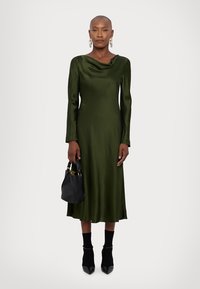 Langere mouwen, olijfgroen jurk van satijnmateriaal met een buiklijn. Model houdt een zwarte tas vast, in combinatie met zwarte sokachtige schoenen.