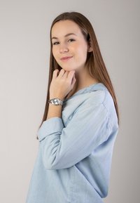 Silberne Armbanduhr an einer Hand, die ein hellblaues Langarmhemd trägt. Weicher, strukturierter Stoff mit einem aufgerollten Ärmeldesign. Neutraler Hintergrund.