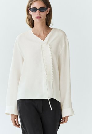 Femme portant des lunettes de soleil noires, un chemisier blanc à manches longues avec un détail de volants verticaux et un pantalon noir sur un fond clair uni.