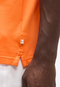 Κοντινό πλάνο ενός σκουρόχρωμου βραχίονα δίπλα στο τελείωμα ενός πορτοκαλί πουκαμίσου με μια μικρή λευκή ετικέτα «ck» και λευκά παντελόνια.