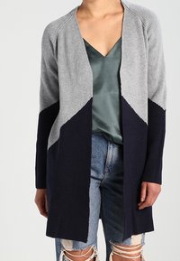 Grau-navy gerippte Strickjacke mit offenem Frontdesign, langen Ärmeln und diagonaler Farbblockgestaltung, getragen über einem satin-grünen Oberteil.