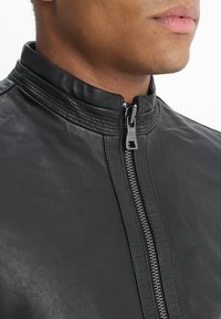 Close-up van het onderste deel van het gezicht en de nek van een persoon die een zwart leren jack met een opstaande kraag en ritssluiting draagt.