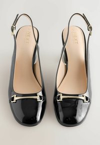 Musta patentoitu nahka slingback-kengät kiiltävällä pinnalla ja kultainen koristeperävarsi varvasalueen yli, vaaleaa taustaa vasten.