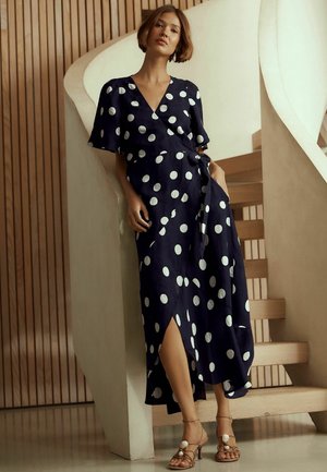 Woman in navy blue wrap dress with white polka dots standing by curved staircase, wearing beige strappy heels.