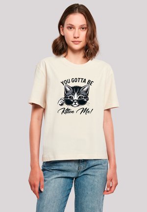 Cremefarbener Baumwoll-T-Shirt mit einem Grafik eines Katers mit Brille und dem Text "DU MUSST MICH BEMÄNGELN!" in fettem schwarzen Schriftzug.