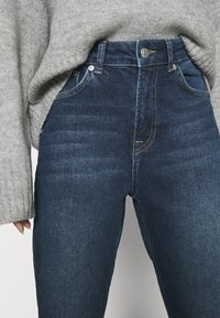 Jeans a vita alta di colore blu scuro con chiusura a bottone, indossati con un maglione grigio lavorato a maglia parzialmente infilato dentro.