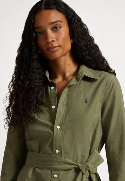 Olivo zelena srajca s sponkami iz mehke tkanine, z klasičnim ovratnikom, dolgimi rokavi in zavezanim pasom. Vključuje majhen temnomoden logotip.