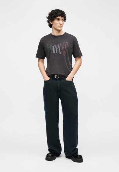 Νέος με σγουρά μαλλιά που φοράει ξεθωριασμένο μαύρο t-shirt με το λογότυπο "KOOPLES", φαρδιά σκούρα τζιν, ζώνη με σχέδια και μαύρα χοντρά παπούτσια, με τα χέρια στις τσέπες.