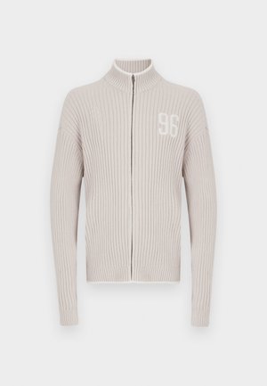 Jasnobeżowy dzianinowy sweter na suwak z prążkowaną fakturą, wysokim kołnierzem oraz białym emblematem "96" na lewej piersi. Długie rękawy i dopasowany dół.