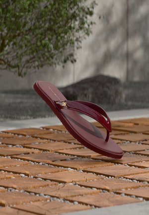 Βουργουνδί παντόφλα με γυαλιστερές λουράκια και χρυσό λογότυπο, αιωρούμενη πάνω από ξύλινα πλακάκια με μοτίβα σε εξωτερικό χώρο.