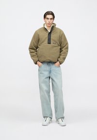 Fiatal férfi olívazöld polárpulóverben, világoskék farmerben és fehér sportcipőben, zsebre dugott kezekkel áll fehér háttér előtt.