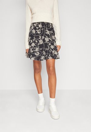 Femme portant une mini-jupe noire à motif floral, un pull crème, des chaussettes blanches et des baskets blanches, debout devant un fond uni.