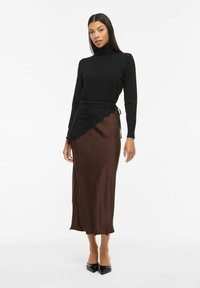 Femme portant un pull à col roulé noir, une jupe midi en satin marron avec un par-dessus en dentelle noire et des escarpins noirs à bout pointu, sur fond blanc.