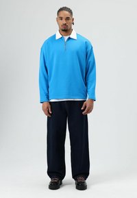 Blauwe sweatvest met een witte kraag, ritssluiting aan de voorkant en lange mouwen, gecombineerd met donkerblauwe wijde jeans en zwarte schoenen met roze veters.