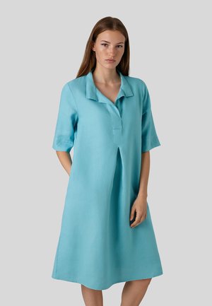 Frau stehend in einem lockeren, knielangen, hellblauen Kleid mit Kragen und halblangen Ärmeln vor einem schlichten grauen Hintergrund.