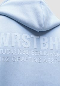 Lyseblå hoodie lavet af blødt stof, med hævet hvid tekst: "WRSTBHVR STUDIO (030) BERLIN MON. 102 CRAFTING AESTHETICS."