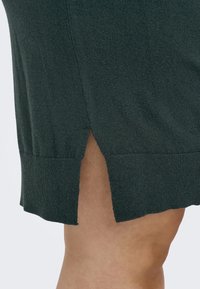 Jupe tricotée vert foncé avec une finition texturée, dotée d'une fente sur le côté et de détails en côtes à l'ourlet. Le tissu semble doux et léger.