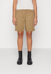 Corti di cotone color khaki con orli arrotolati, dotati di una tasca aperta e una consistenza liscia, abbinati a stivaletti neri alla caviglia.