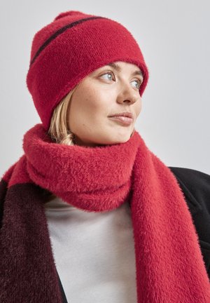 Bonnet tricoté rouge et duveteux avec un accent en rayures noires ; écharpe épaisse assortie dans une texture et une teinte similaires ; portée avec un haut blanc.