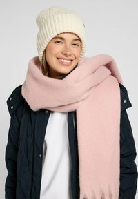 Sciarpa di lana rosa chiaro avvolta attorno a una giacca nautica trapuntata, abbinata a un berretto a coste beige. Texture liscia e aspetto caldo.