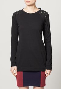 Maglietta nera a maniche lunghe con maniche raglan, caratterizzata da borchie verdi sulle spalle e un orlo testurizzato. Materiale in cotone morbido.