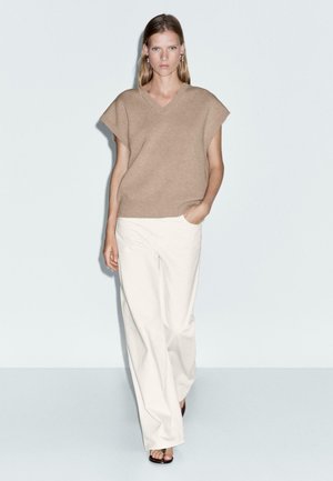 Donna che cammina indossando un maglione senza maniche beige a scollo a V, pantaloni bianchi a gamba larga, orecchini a cerchio argento e tacchi aperti neri su uno sfondo semplice.