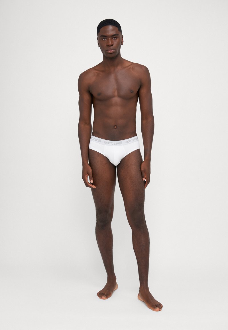 Caleçons blancs en coton avec une ceinture grise arborant le logo "Roberto Cavalli". Texture lisse et coupe ajustée, modèle debout pieds nus.