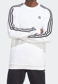 Mężczyzna w białej bluzie Adidas z czarnym wzorem trzech pasków na rękawach i małym logo Adidas na piersi.