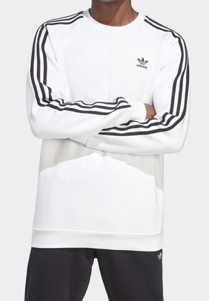 Man wearing white Adidas sweatshirt with black three-stripe design on sleeves and small Adidas logo on the chest.