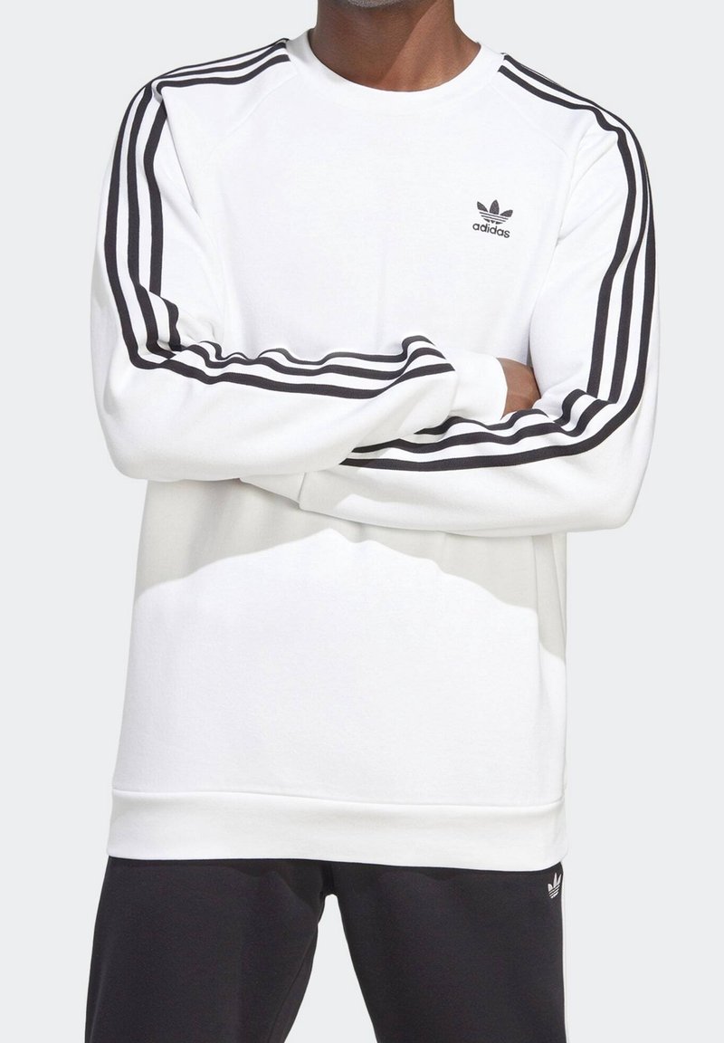 Mężczyzna w białej bluzie Adidas z czarnym wzorem trzech pasków na rękawach i małym logo Adidas na piersi.
