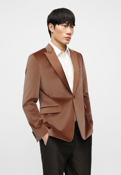 Καφέ σατέν σακάκι με γυριστό πέτο, μονόπετο σχέδιο και δύο μπροστινές τσέπες, συνδυασμένο με λευκό πουκάμισο και σκουρόχρωμο παντελόνι με σχέδια.